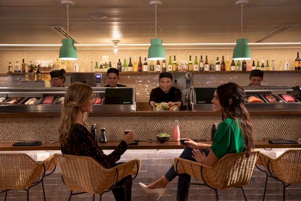 Two people sit at a sushi bar, chatting and having drinks. Chefs behind the counter prepare sushi. Bottles line the background shelf.