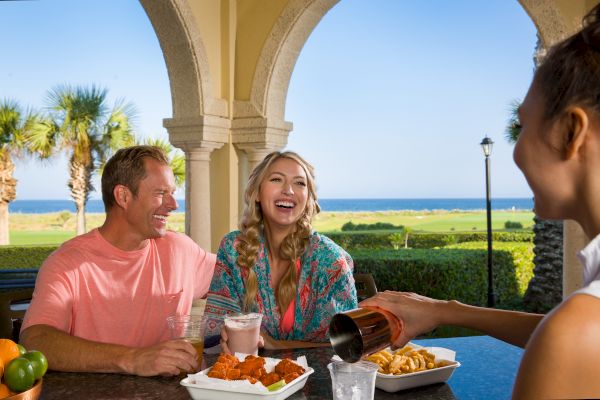 A group enjoying drinks and food outdoors by the ocean, with palm trees and clear skies in the background.