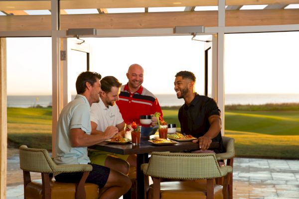 A group of four men are laughing and enjoying a meal together in a bright room with an outdoor view.