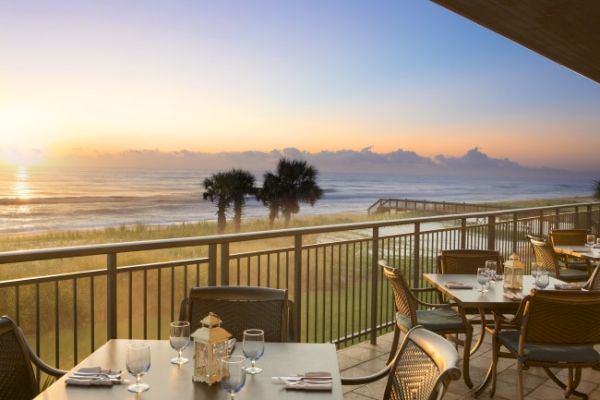 A scenic beachfront dining area with set tables, overlooking the ocean and palm trees at sunrise or sunset.