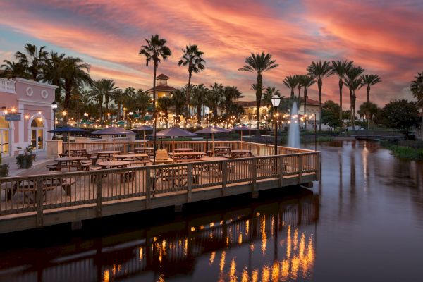A picturesque waterfront dining area with palm trees, string lights, and a colorful sunset sky reflected in the calm water.
