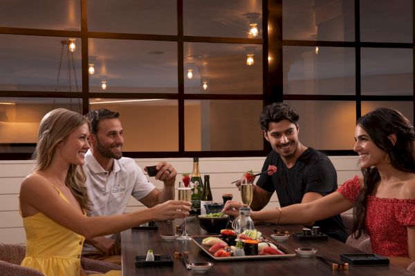 Four people enjoying sushi and drinks at a restaurant, smiling and engaging in conversation in a warmly lit setting.