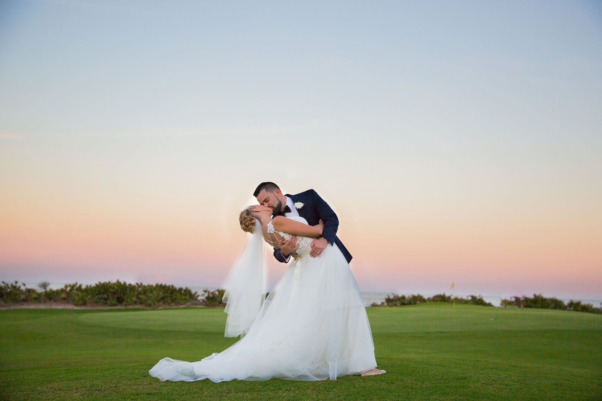 A couple in wedding attire shares a romantic kiss at sunset on a grassy field.