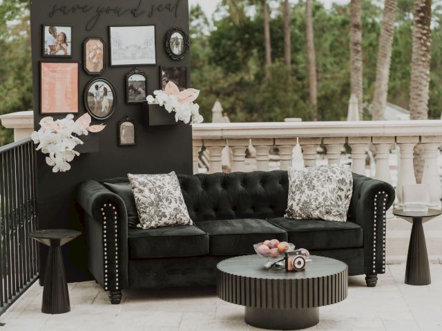 A black couch with patterned pillows sits outdoors, surrounded by tables. A decorated wall with photos and flowers is behind it.