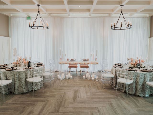 An elegant, softly-lit dining setup with round tables, floral centerpieces, and candles, featuring white drapes and chandeliers.