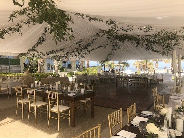 An elegant outdoor event setup with tables, gold chairs, and greenery under a white draped tent, near a water view.