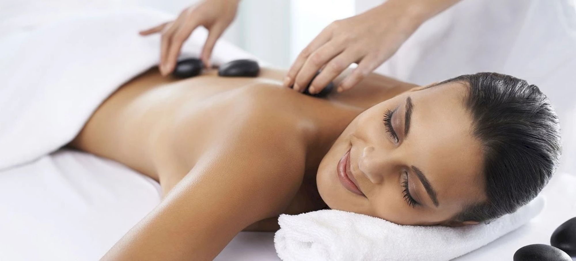 A person receives a relaxing hot stone massage while lying on a massage table, covered partially by a towel, with a smile of contentment.