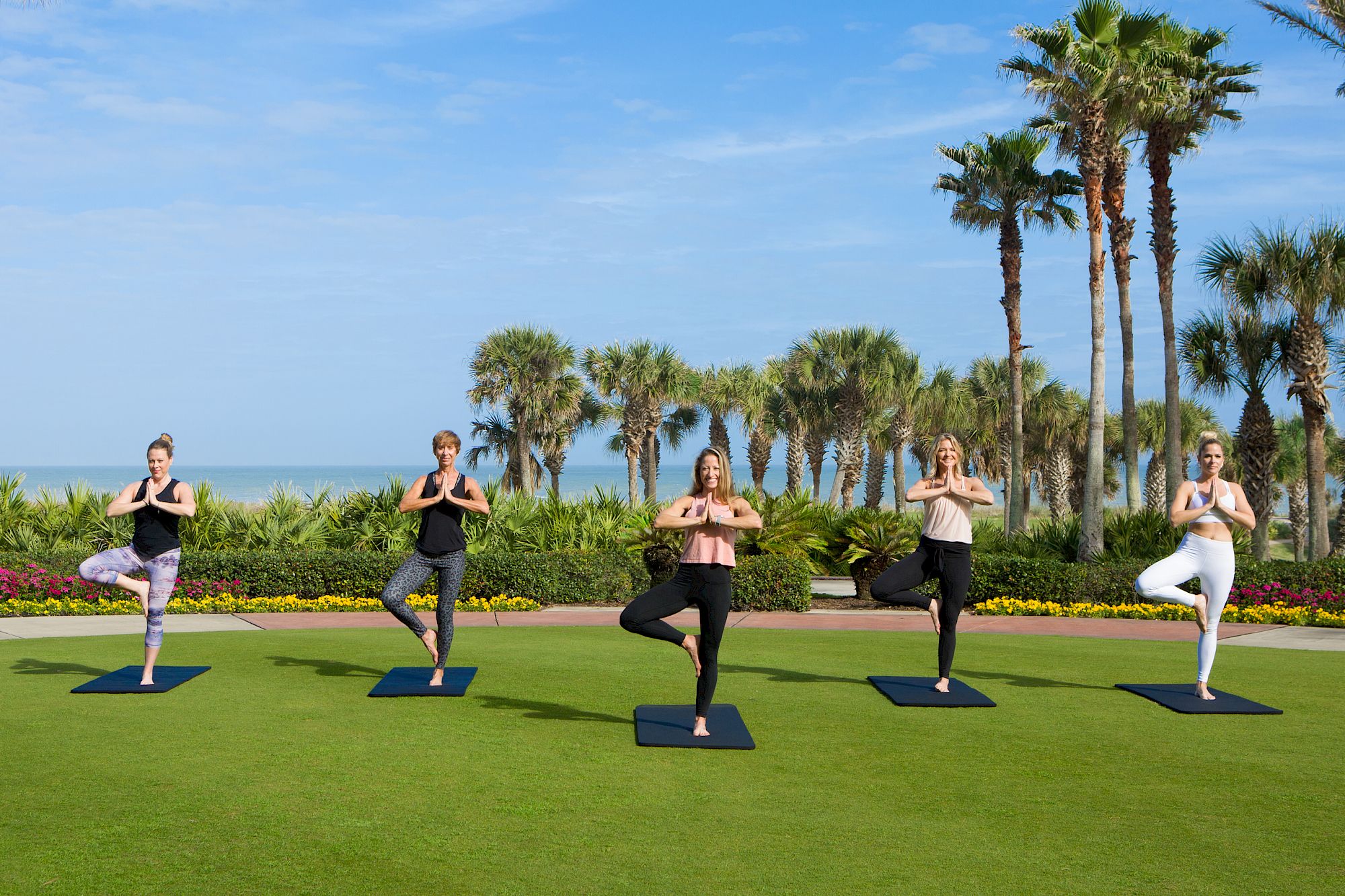 A group of five people doing yoga outdoors on mats, surrounded by palm trees and a clear blue sky.