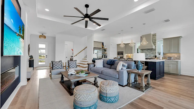 A modern living room with a sectional sofa, TV, ceiling fan, and open kitchen with pendant lights and island.