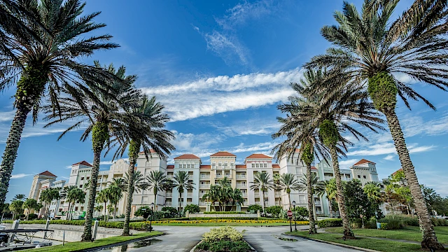 A large building with multiple balconies, surrounded by palm trees and clear skies, viewed from a road entrance.