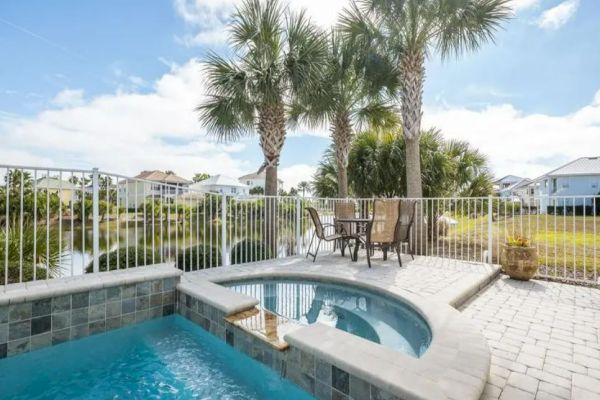 A serene patio with a pool and jacuzzi, surrounded by palm trees and a railing, overlooking a pond, and featuring a patio set.
