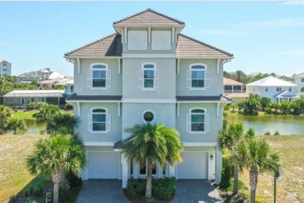 A large three-story house with a tiled roof, surrounded by palm trees, near a body of water.