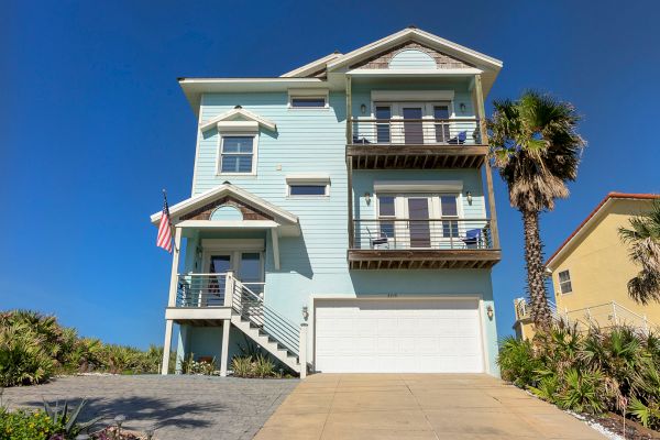 A light blue seaside house with balconies, an American flag, garage, and a palm tree in front under a clear blue sky.