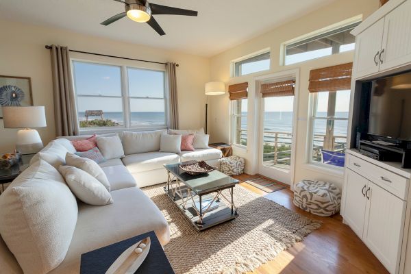 A cozy living room with a white sectional sofa, ocean view, and a TV cabinet with wooden floors complementing the bright space.