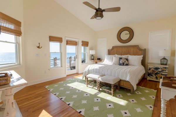 A cozy bedroom with a large bed, sea view through windows, wooden floors, decorative elements, and a light green patterned rug.
