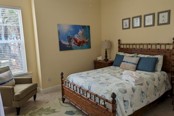 A cozy bedroom with a wooden bed, ocean-themed bedding, and coastal artwork on the walls, featuring a chair and a large window.