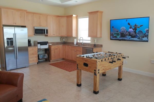 A kitchen and lounge area with wooden cabinets, appliances, a foosball table, and a wall-mounted TV displaying an aquarium scene.