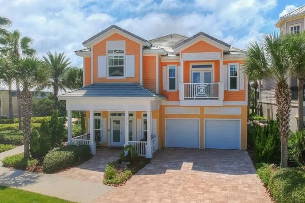 A two-story orange house with white trim, palm trees, and a paved driveway under a blue sky with clouds.