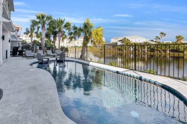 A serene poolside area with lounge chairs, palm trees, and a view of a fenced waterway in a residential setting.