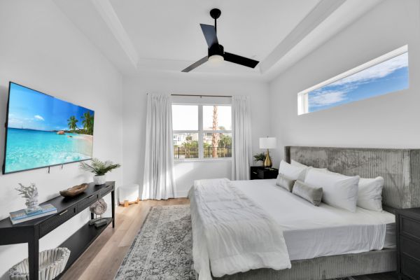 A modern bedroom with a large bed, a wall-mounted TV, a ceiling fan, and a window view, decorated in neutral tones.