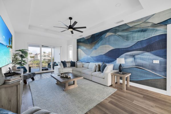 A modern living room with a large white sectional, abstract blue wall art, ceiling fan, TV, and view of a balcony outside the window.