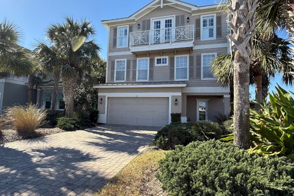 A two-story house with a garage, surrounded by palm trees and landscaped greenery, under a clear blue sky.