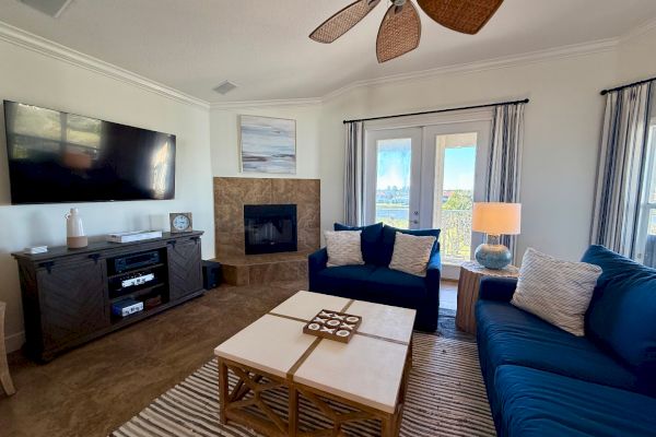 The image shows a modern living room with a blue sofa, wall-mounted TV, and fireplace. A ceiling fan, coffee table, and decor complete the setting.