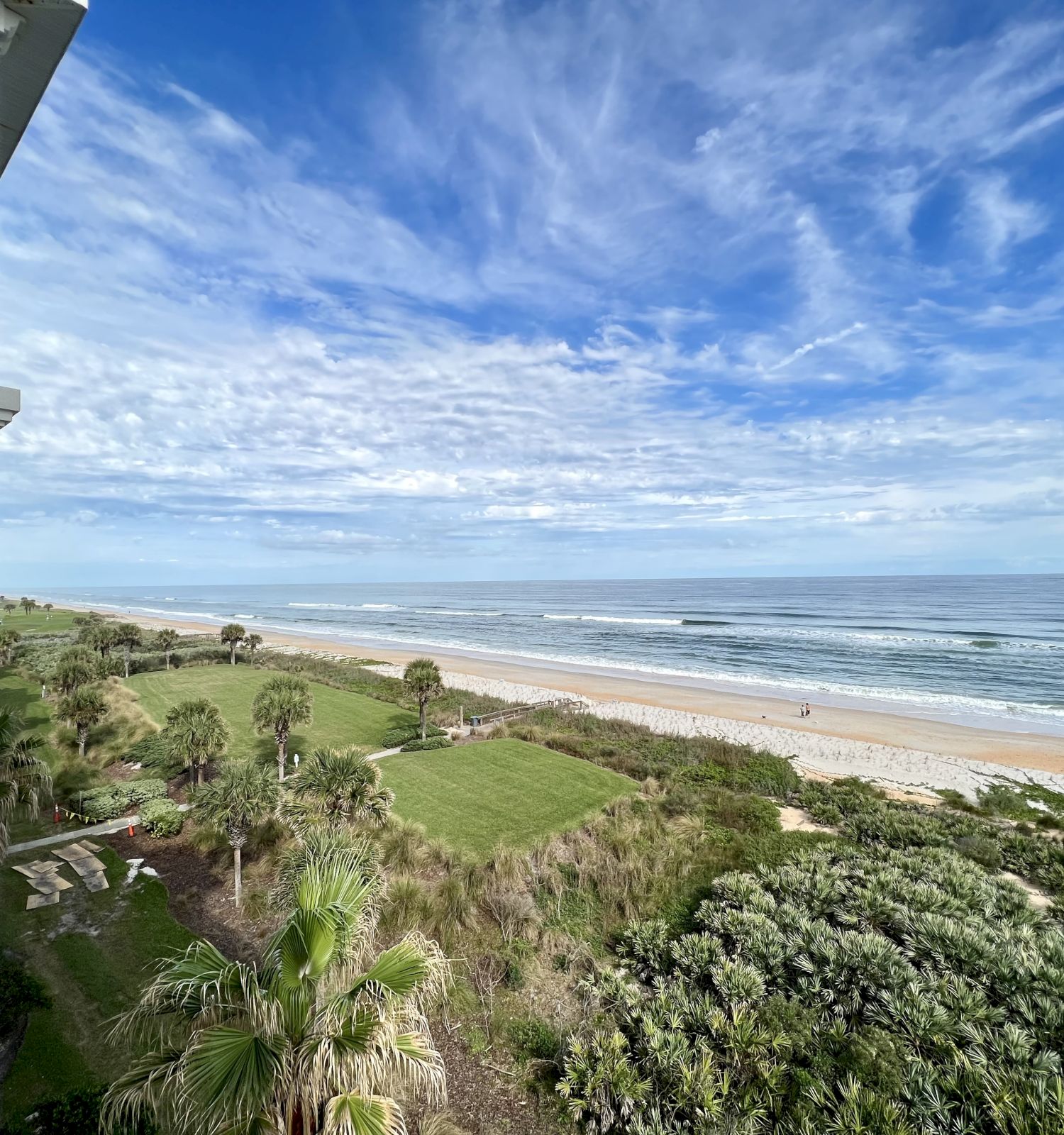 A beach view with a grassy area, palm trees, ocean waves, and a partly cloudy sky, seen from a building balcony.
