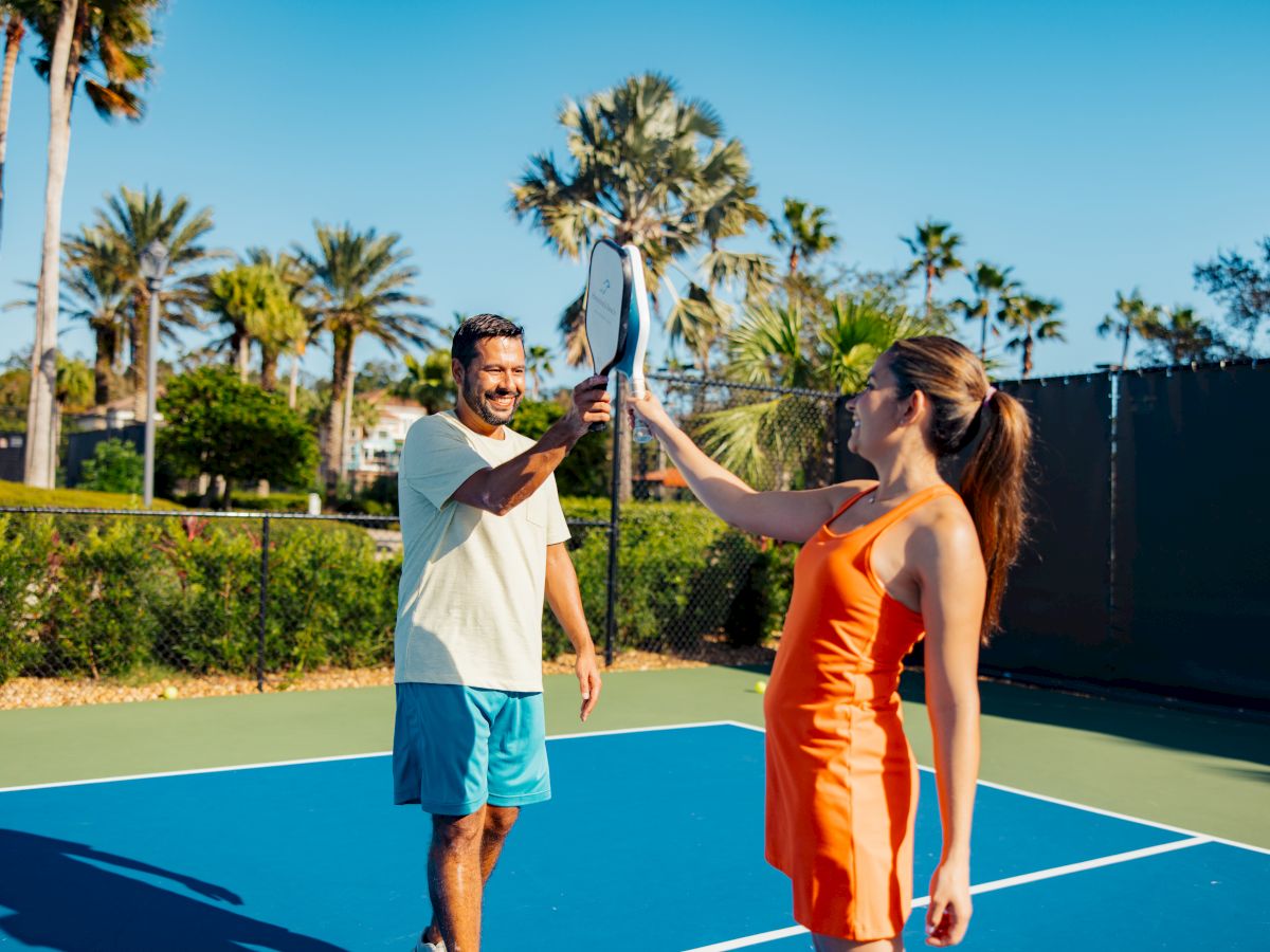 Two people on a tennis court smiling and touching tennis rackets, surrounded by palm trees under a clear blue sky.