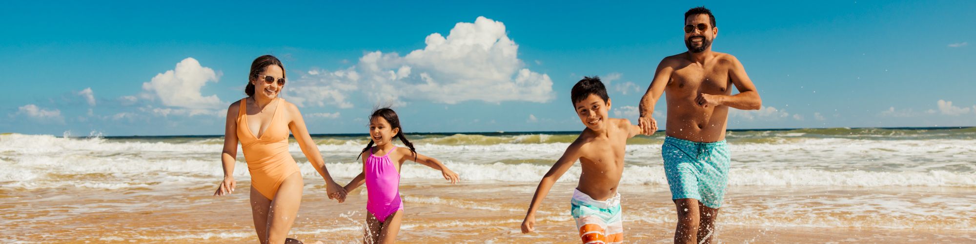 A family of four joyfully runs along the beach, splashing in the waves under a clear blue sky.
