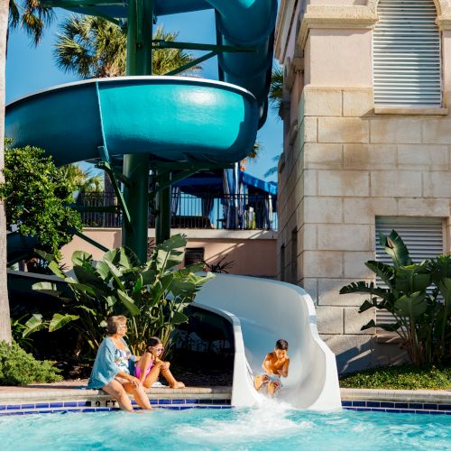 A child slides into a pool from a white slide, with a large blue spiral slide in the background, next to a building.