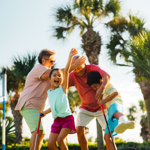 A family of four enjoys a sunny day on a golf course, laughing and celebrating, surrounded by palm trees and lush greenery.