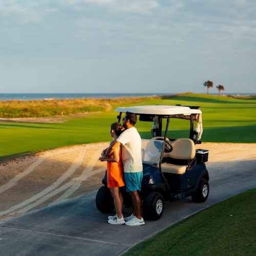 Two people stand beside a golf cart on a golf course path, with a scenic ocean view and blue sky in the background.